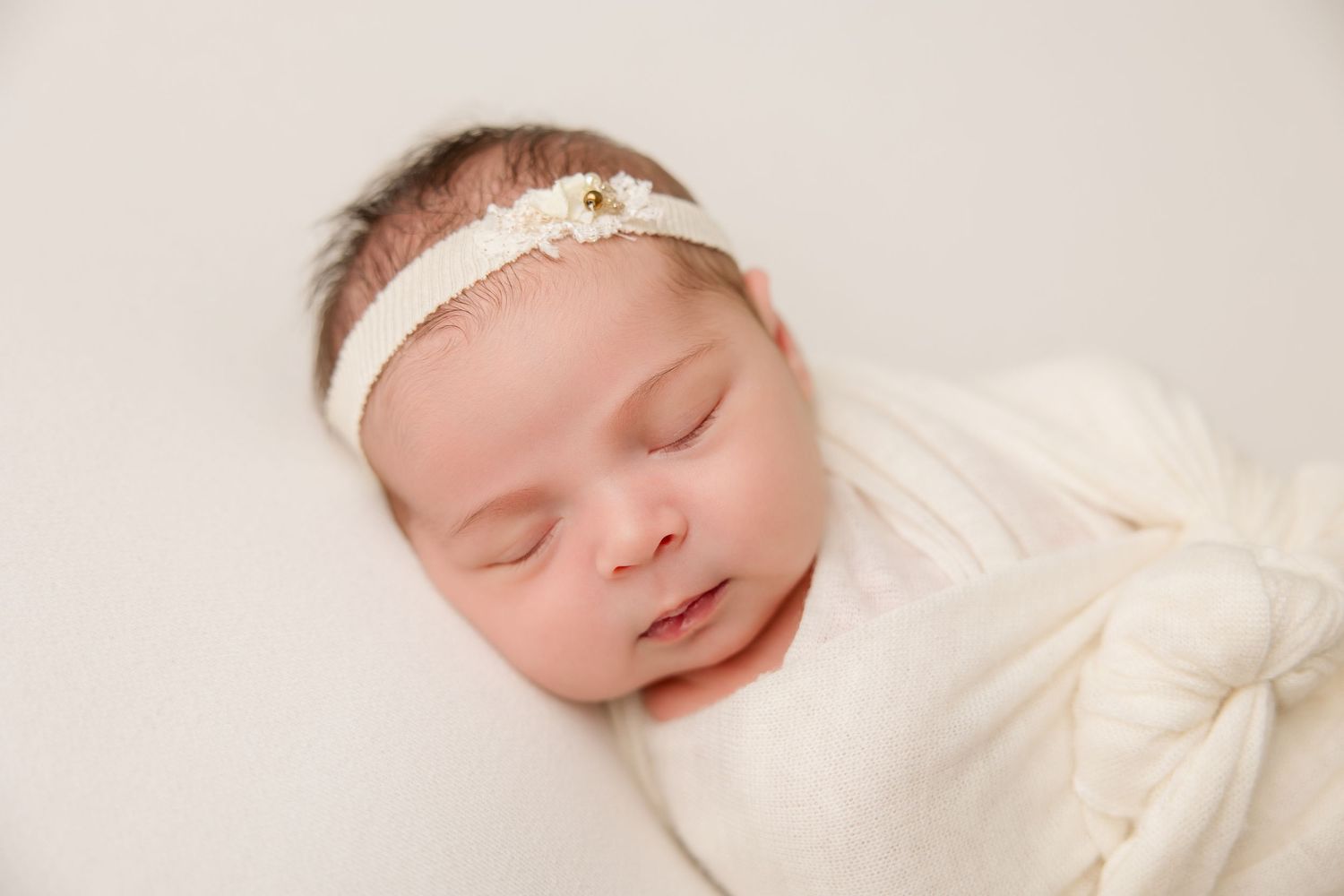 Sleeping newborn wrapped in soft white fabric wearing a delicate lace headband against light background.