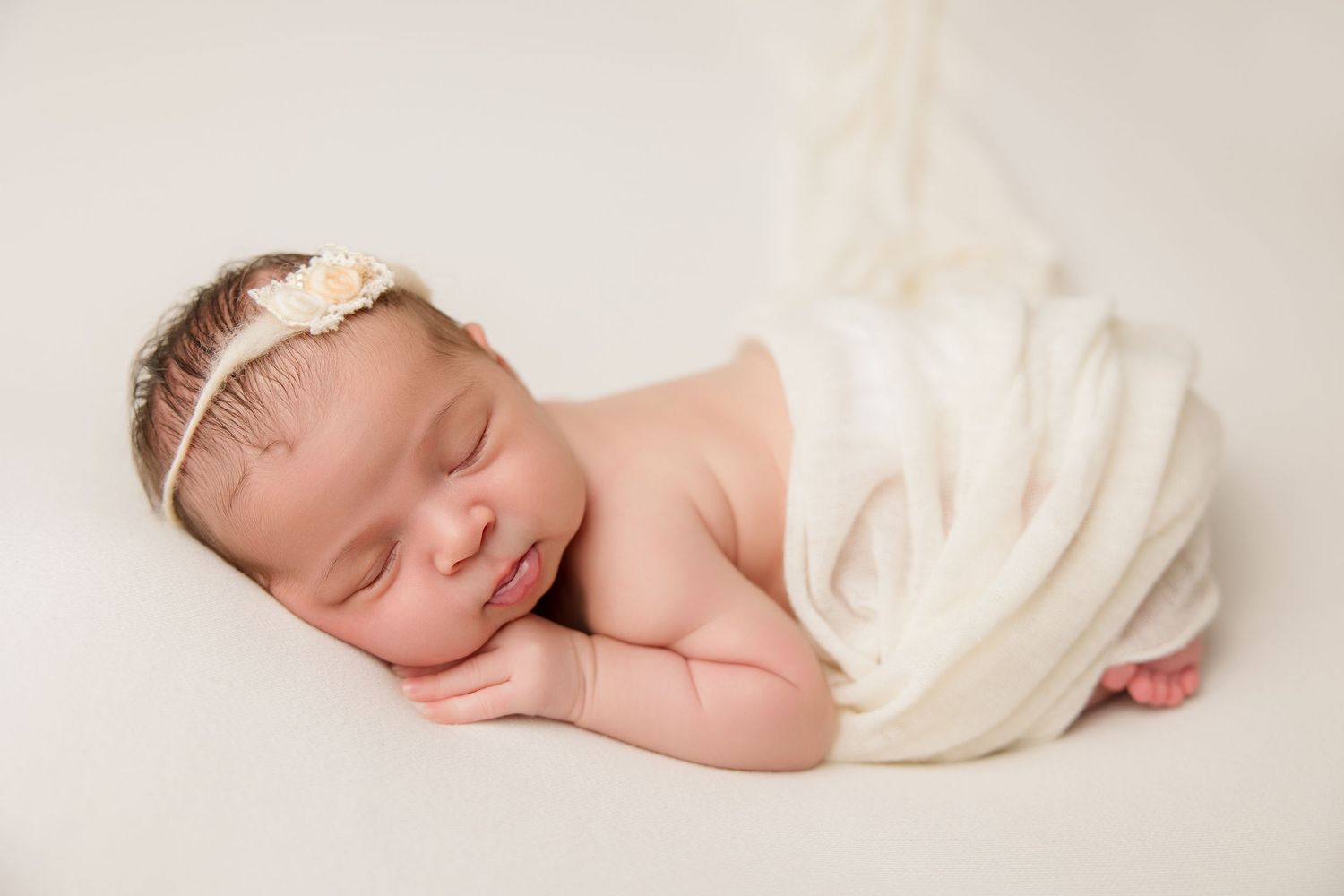 Peaceful newborn sleeping on side wrapped in cream colored fabric with small headband.