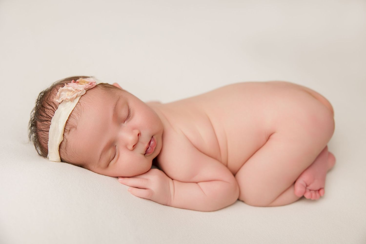 A sleeping newborn baby lies peacefully on soft white fabric in a minimalist studio setting.