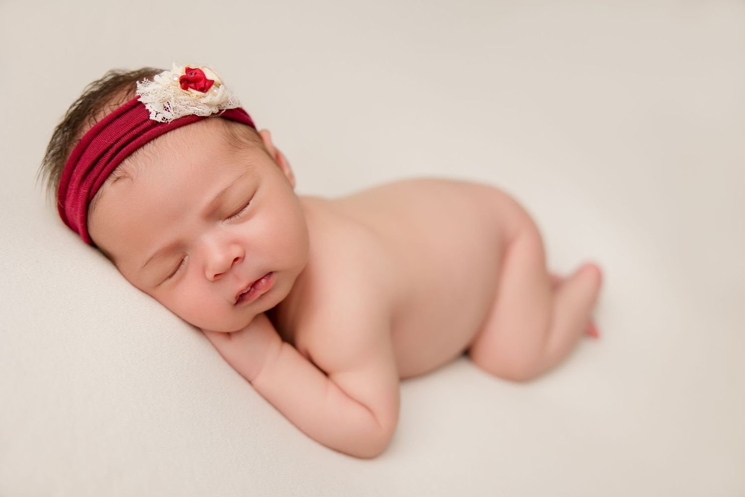 Newborn sleeping on white backdrop wearing red headband with floral accent.