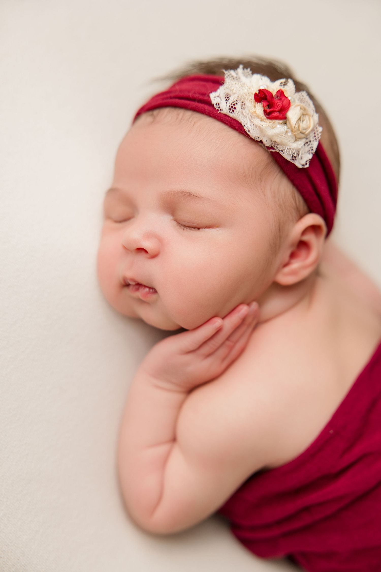 Close-up of sleeping baby with ornate red and white lace headband against light background.