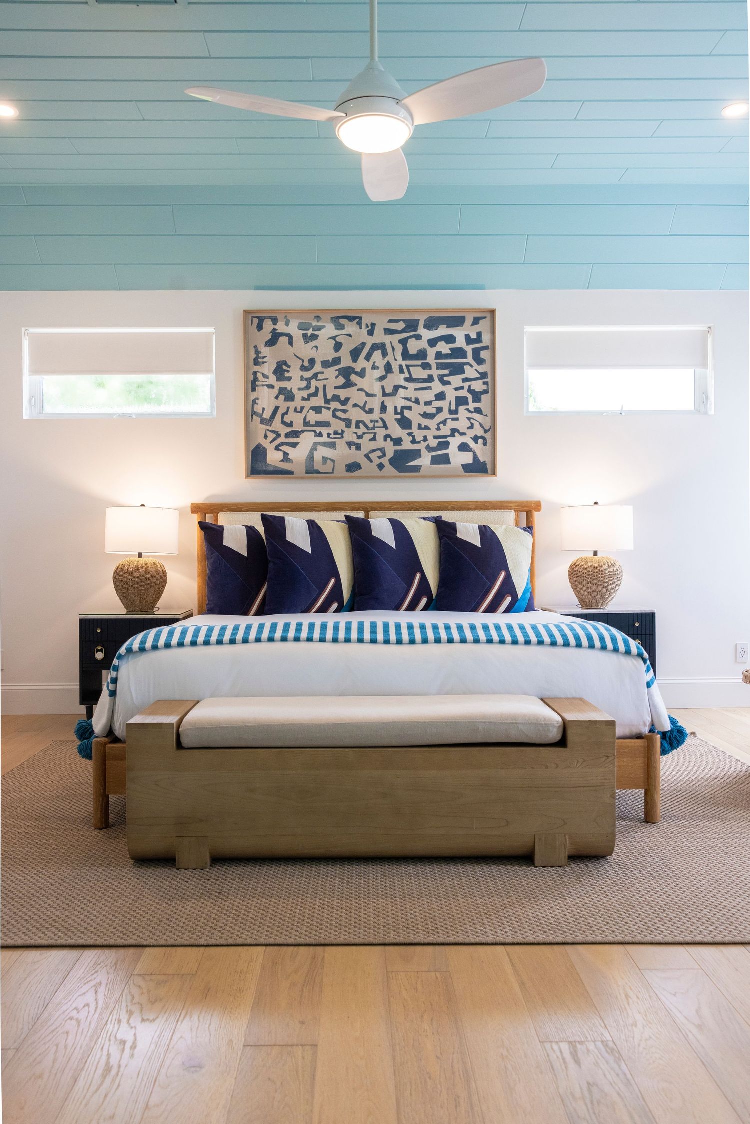 Modern coastal bedroom with blue ceiling, wooden bed frame, navy pillows and abstract wall art between windows.
