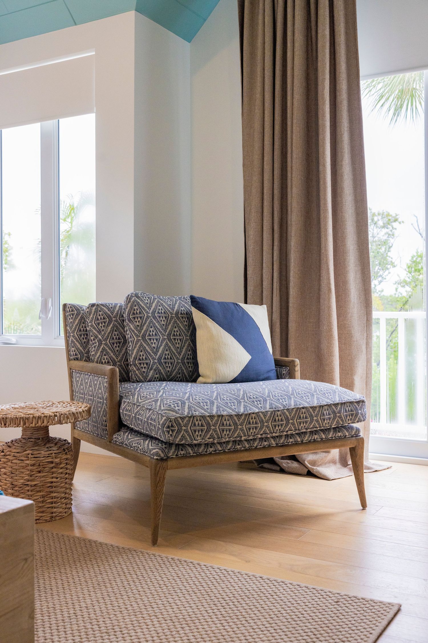 Coastal-style lounge corner with patterned chaise, taupe curtains and hardwood floors beside bright windows.