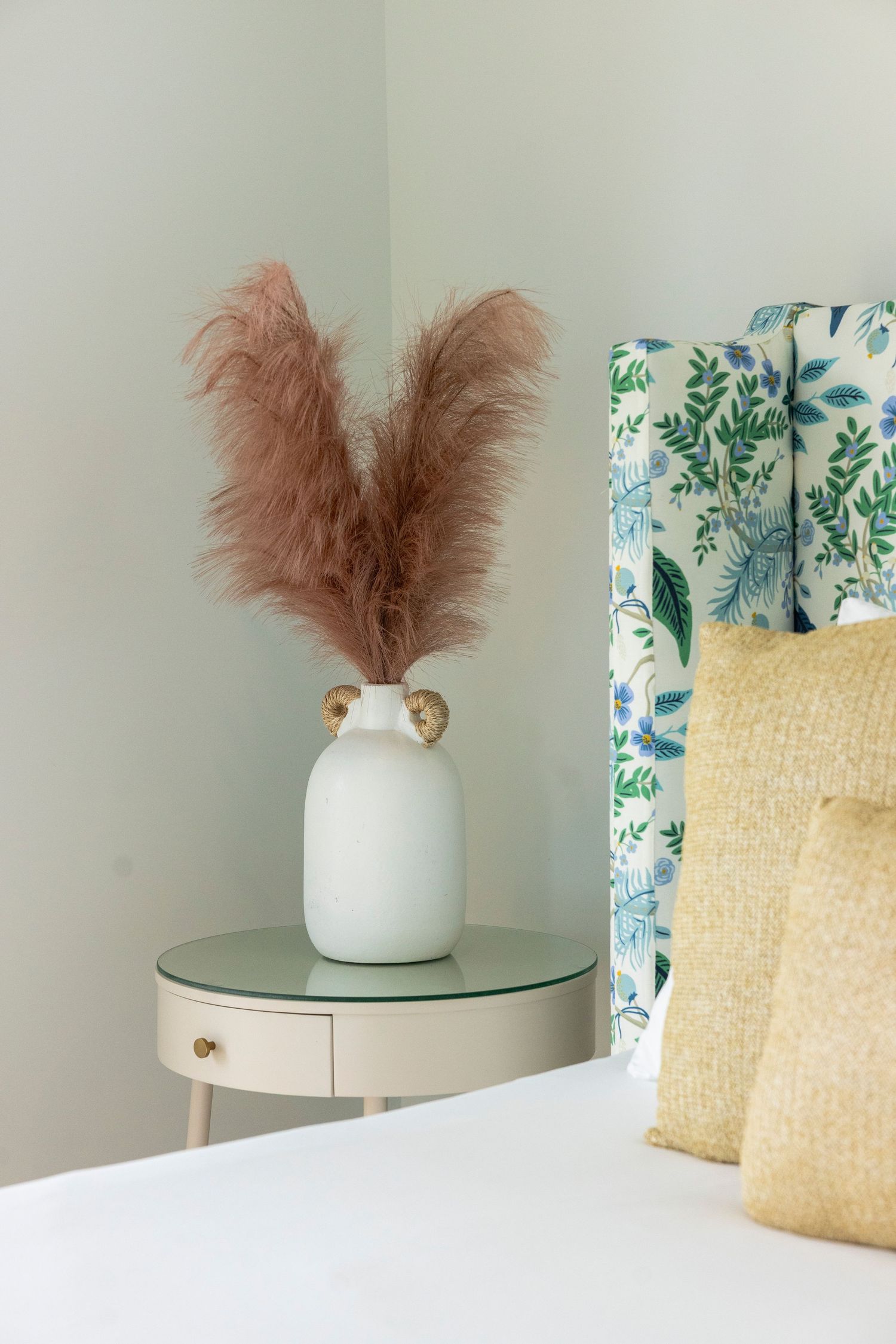White ceramic vase with fluffy pink pampas grass plumes displayed on a round gray side table beside decorative pillows.