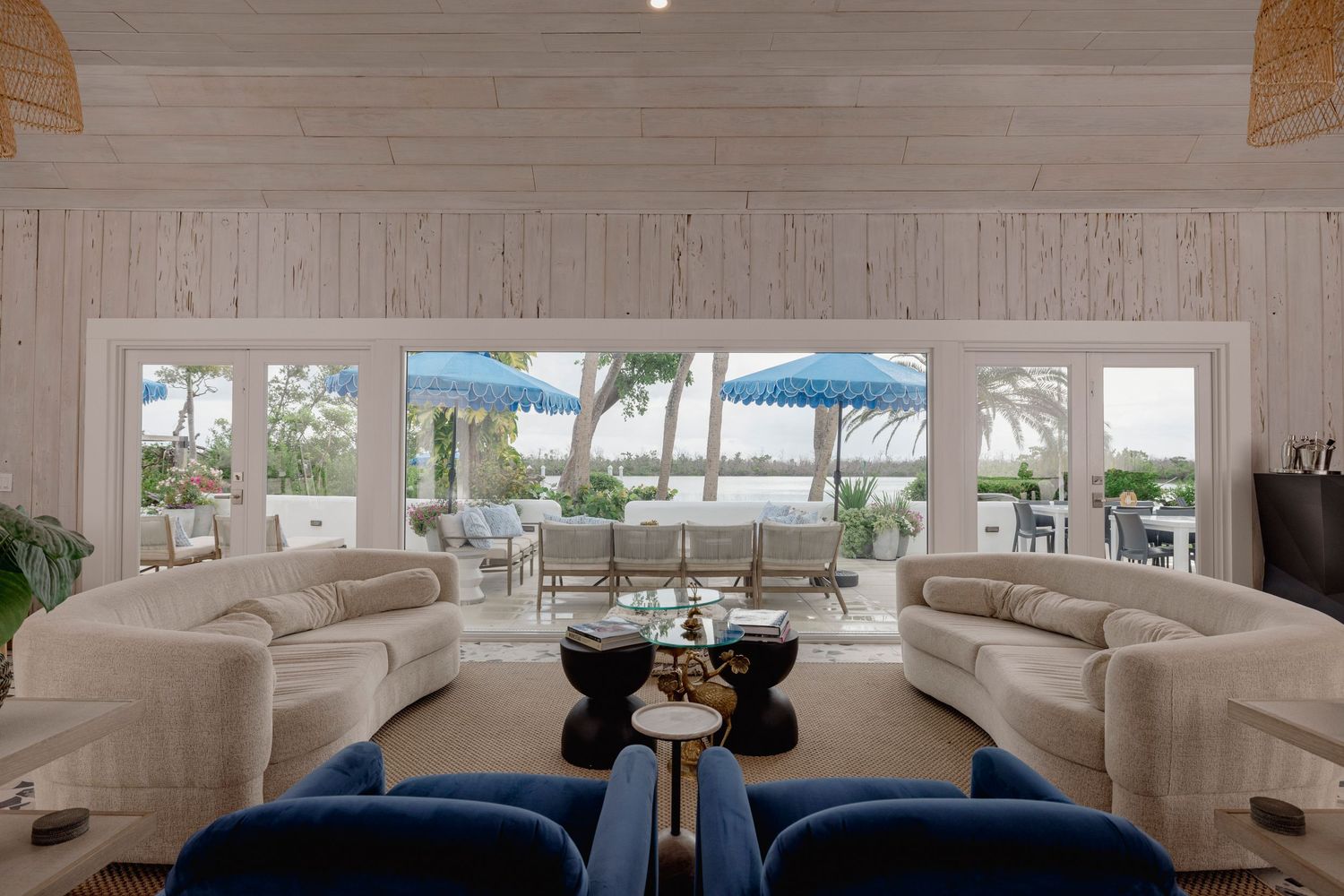 Modern coastal living room features white sofas, light wood beams, and panoramic windows overlooking the water.