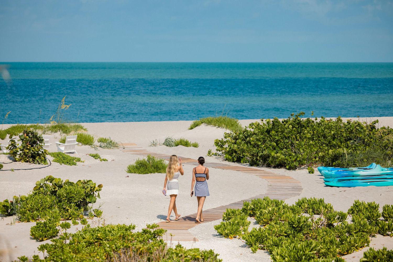 Sandy beach pathway lined with lush greenery leads to turquoise ocean waters under bright blue skies.