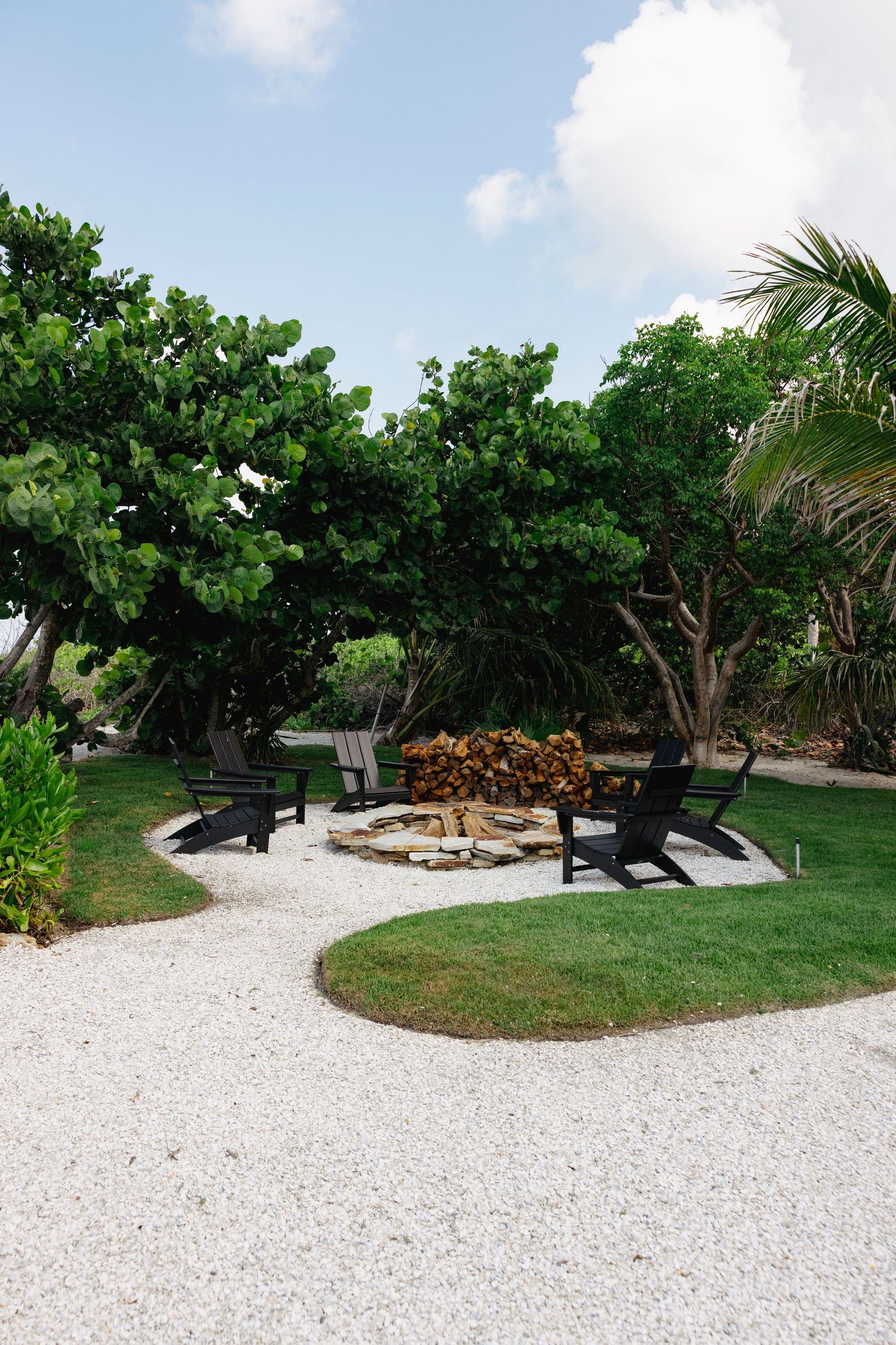 Tranquil outdoor seating area with Adirondack chairs surrounding a fire pit on white gravel path beneath tropical trees.