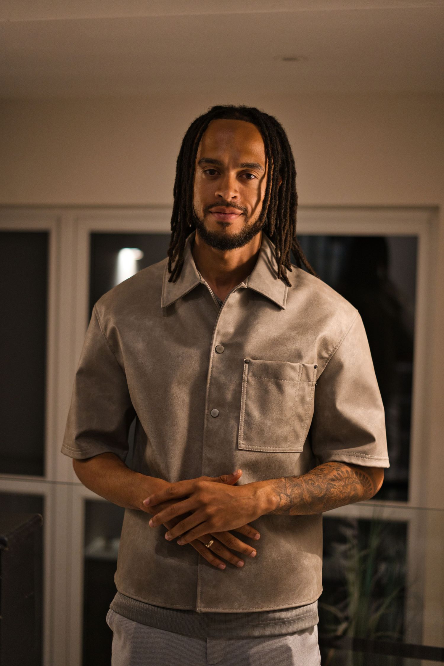 Someone with dreadlocks wearing a light colored shirt against a minimalist interior background.