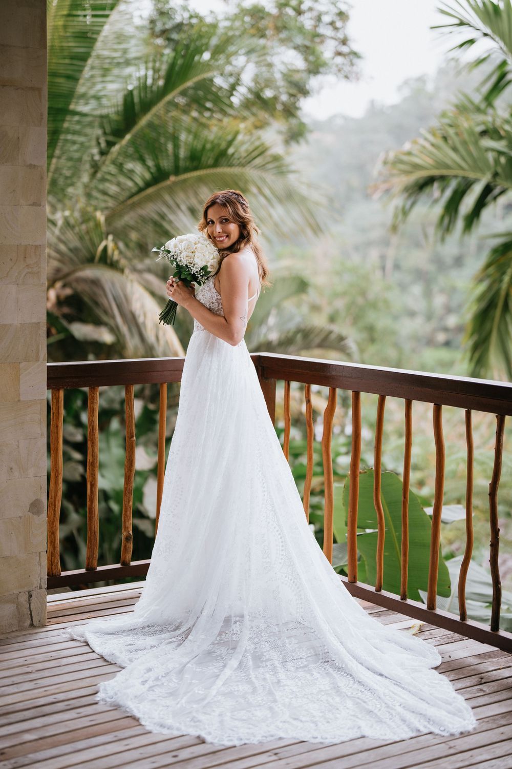 A flowing white wedding dress with lace details displayed on a balcony overlooking tropical scenery.