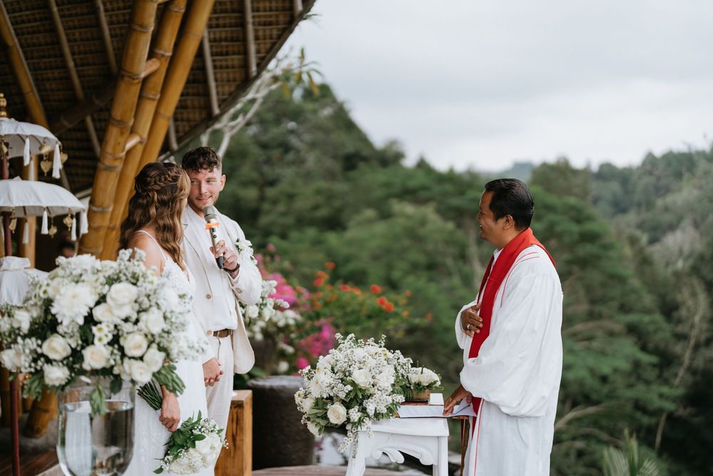 A wedding ceremony in progress under a wooden pavilion decorated with white floral arrangements.