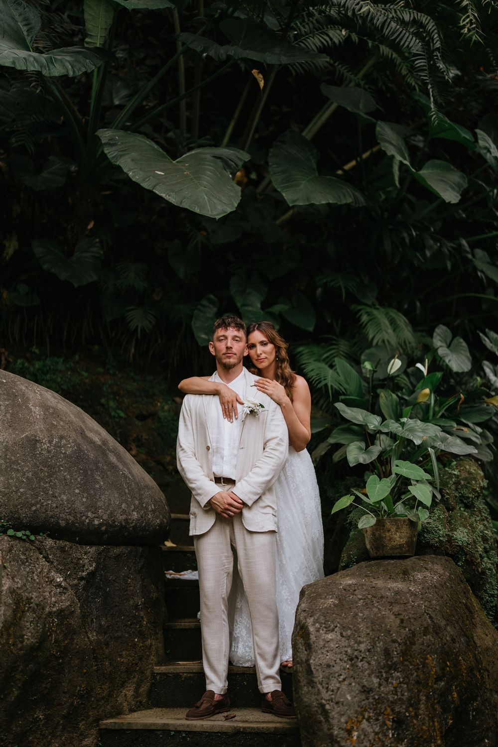 A romantic couple in white outfits pose together on large rocks surrounded by lush tropical foliage.