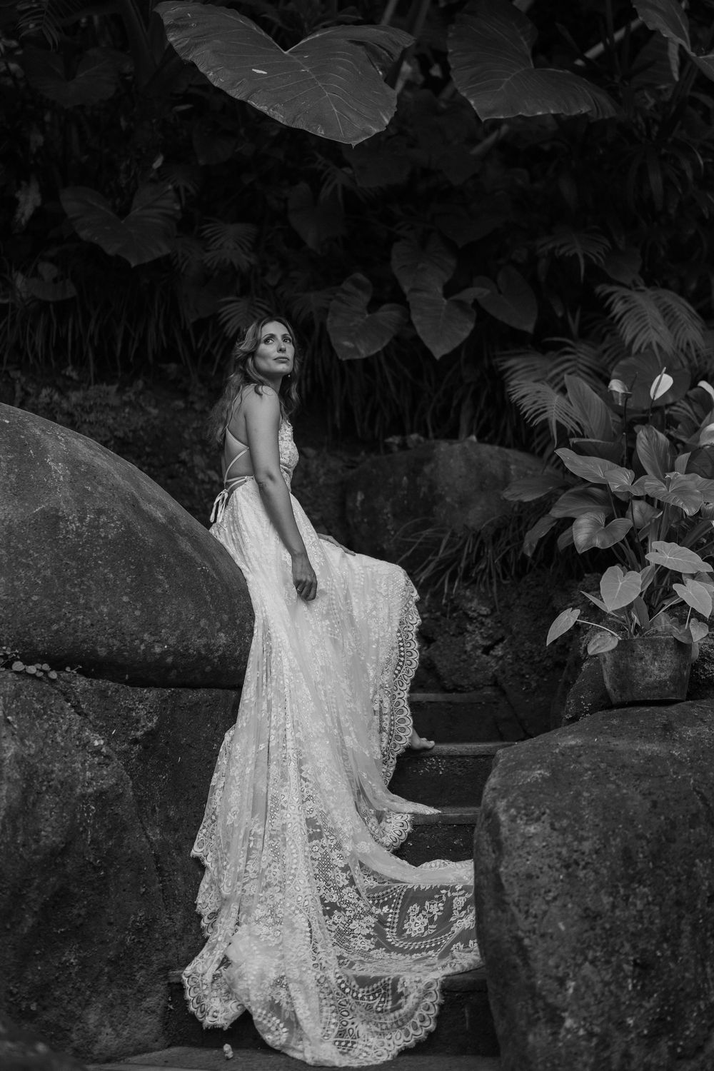 A person in a white lace dress poses gracefully against stone walls in a tropical garden setting.