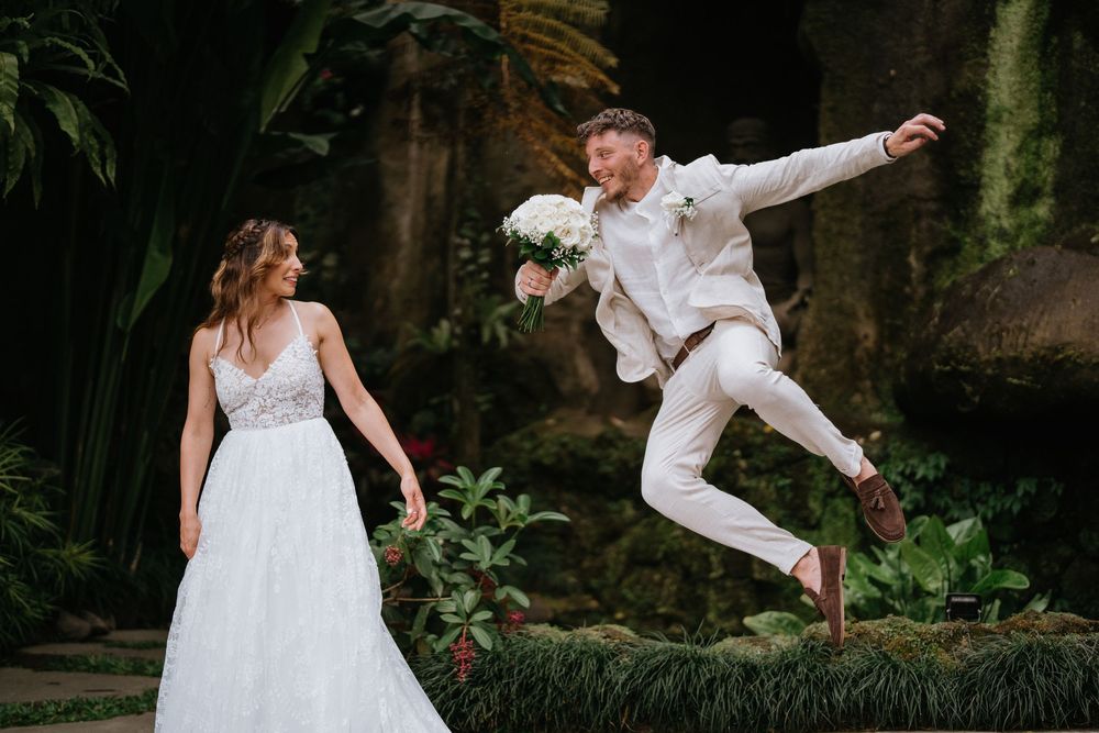 A couple in wedding attire dances and spins playfully in a garden setting.