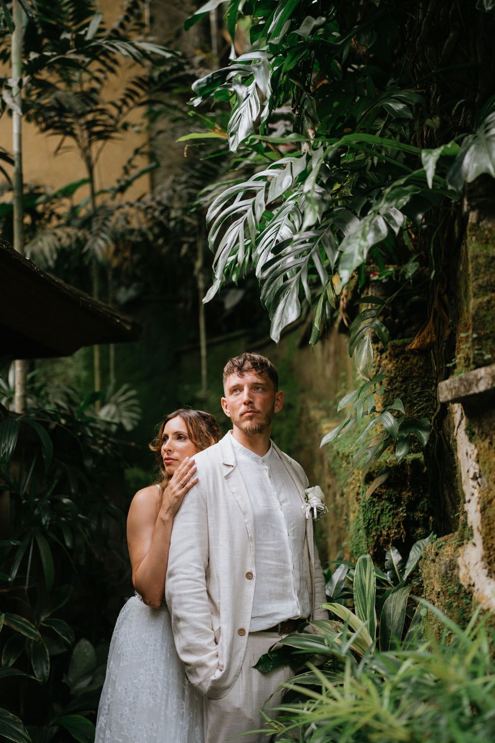 A couple shares an intimate moment among tropical plants and stone walls in white wedding attire.