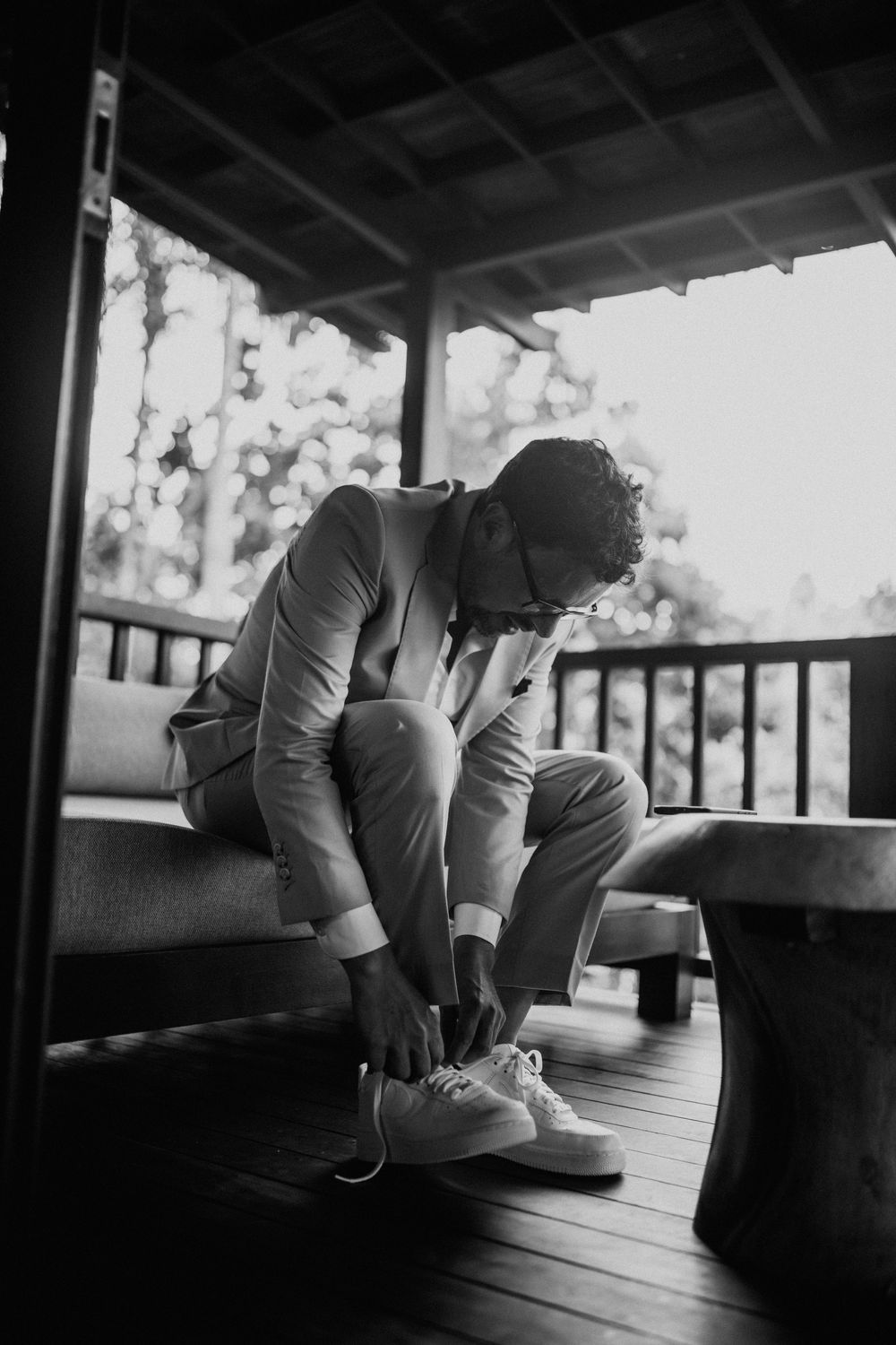 A person in formal attire ties their shoes on a wooden porch in a black and white photograph.