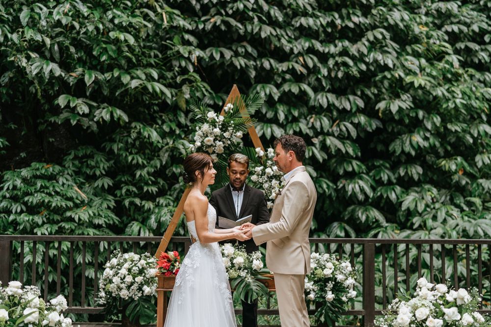 A wedding ceremony takes place on a deck with white floral arrangements against a wall of green leaves.