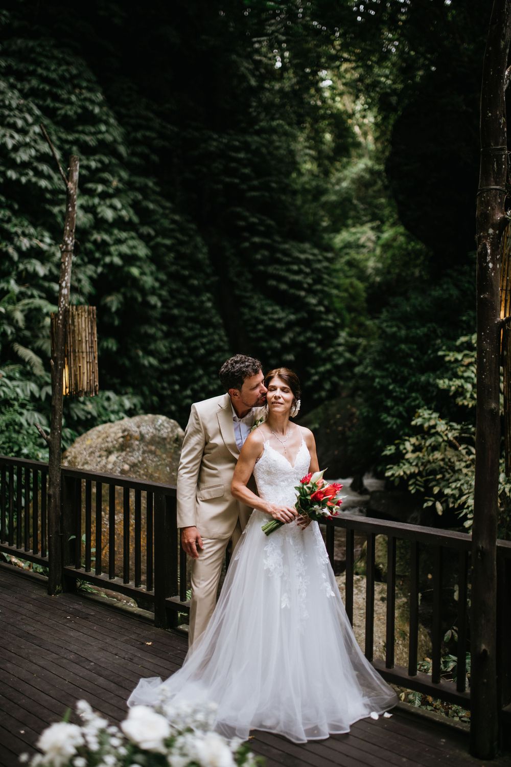 A romantic wedding couple shares an intimate moment on a wooden deck surrounded by lush forest greenery.