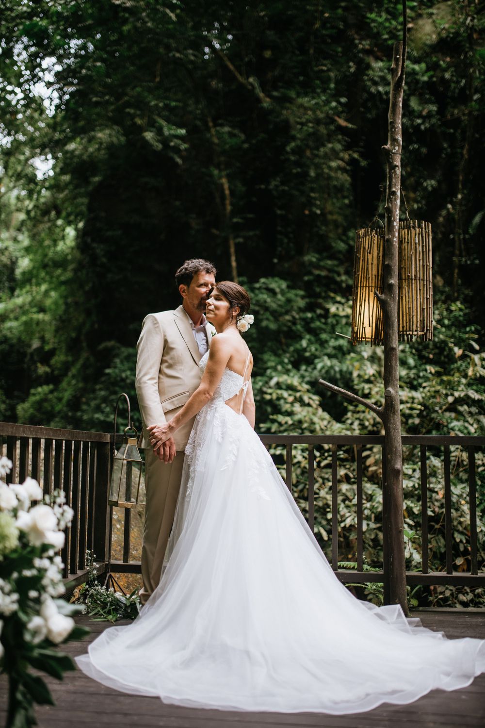 A wedding couple embraces on a rustic wooden deck with hanging lanterns and forest scenery.