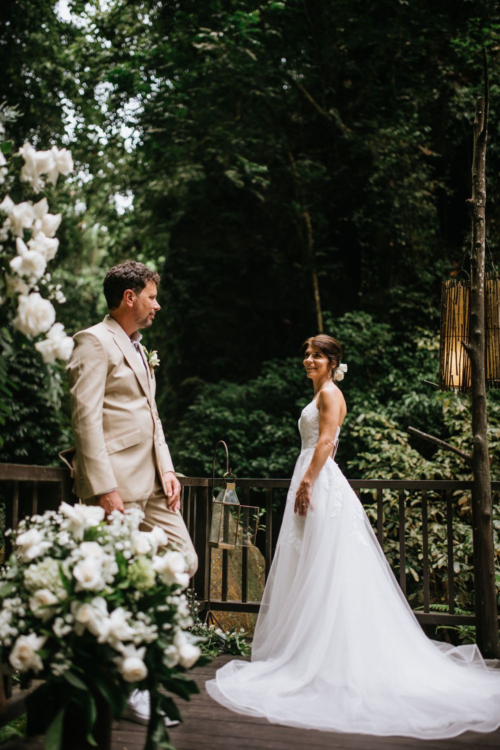 Wedding couple poses elegantly on a wooden deck with white floral decorations in a forest setting.