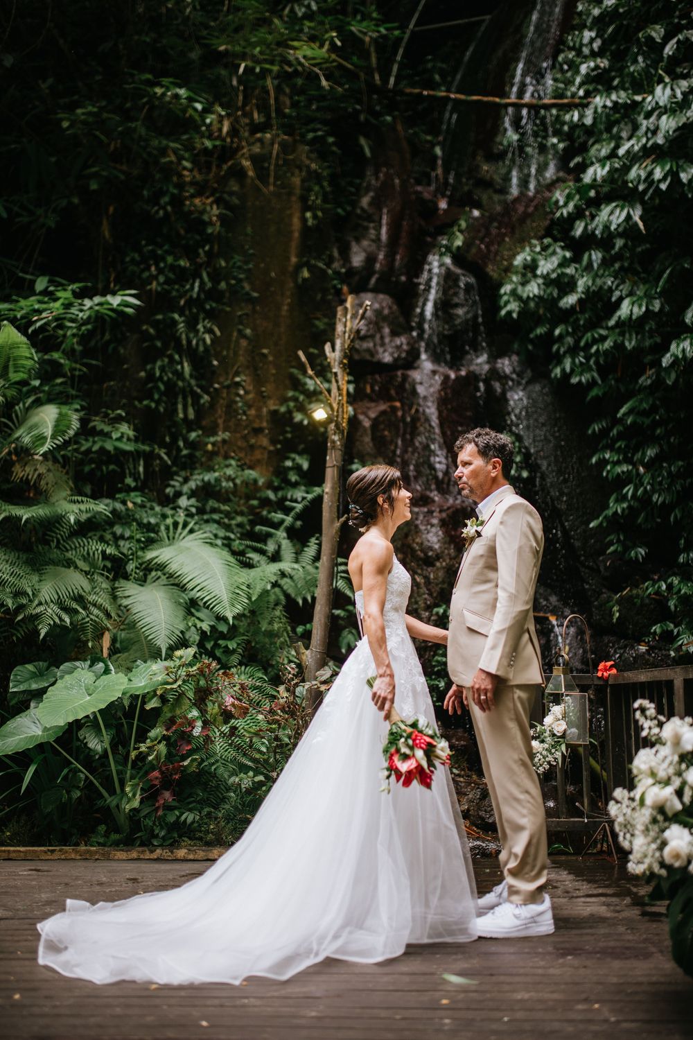 Newlyweds share tender moments on a wooden deck surrounded by tropical foliage and white flowers.