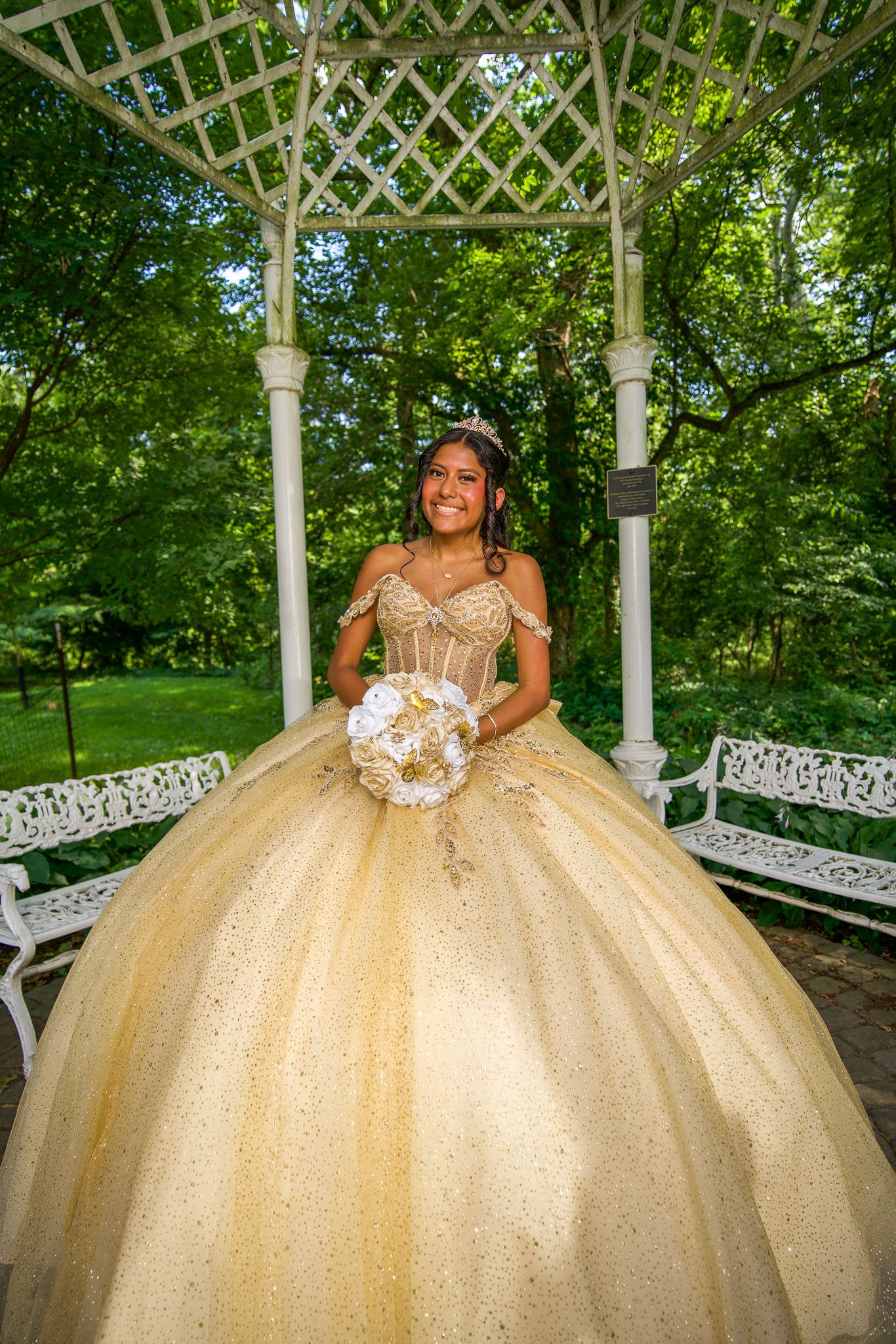QUINCEAÑERA gold ballgown photographed under a white garden gazebo surrounded by lush greenery.