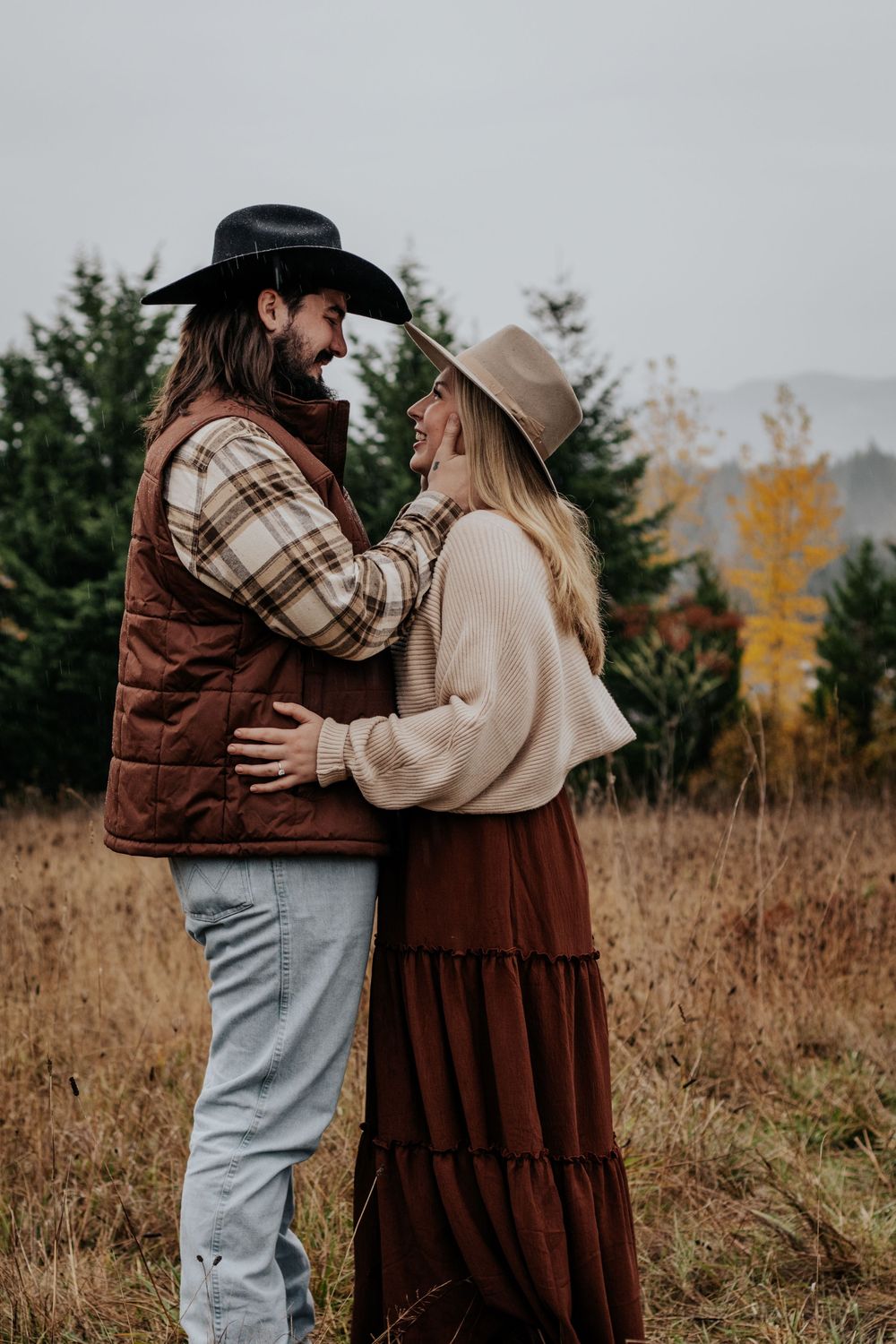 Couple in western hats embracing in autumn field with evergreen trees.