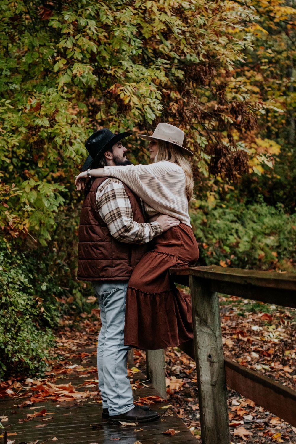 Romantic couple embracing on wooden bridge surrounded by colorful fall foliage, wearing western cowboy hats.