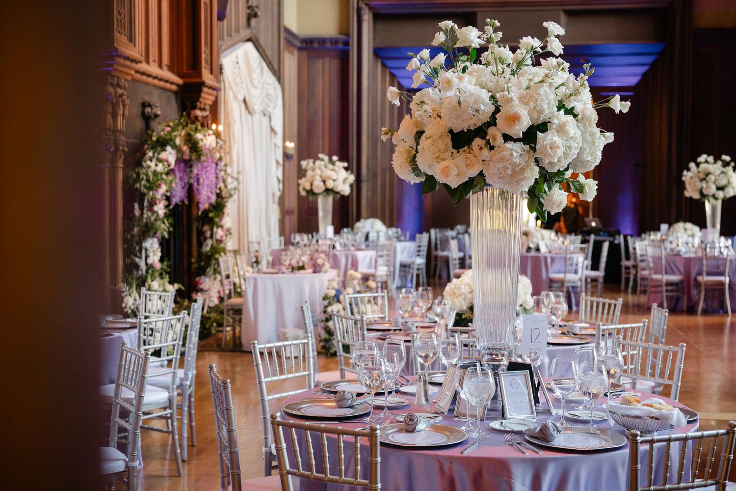 Elegant wedding reception tables decorated with tall white floral centerpieces and candles in a grand ballroom setting.