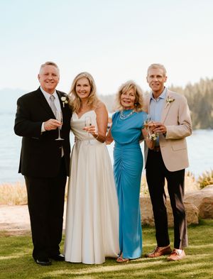 Small wedding party in formal attire poses together on a grassy lawn with scenic water views at sunset.