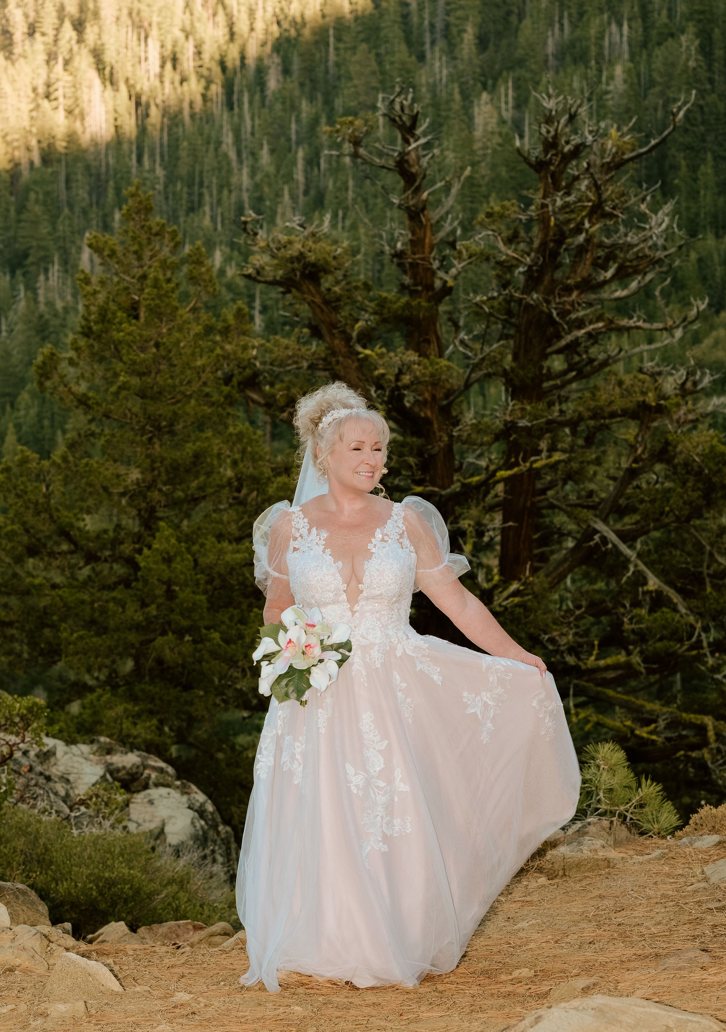 A figure in a flowing white wedding gown poses gracefully among evergreen trees on a mountain trail at sunset.