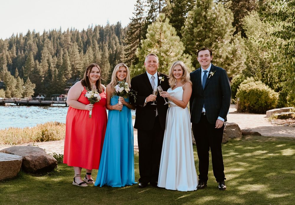 Wedding party wearing red and blue dresses poses together on a grassy lawn by a lake surrounded by evergreen trees.