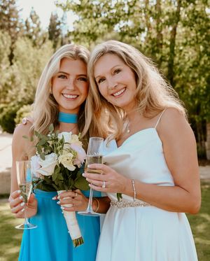 Wedding party friends sharing celebratory drinks and laughter in outdoor garden setting wearing blue and white dresses.