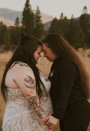 A couple in formal attire embraces in a mountain field at sunset with mountains and trees in the background.
