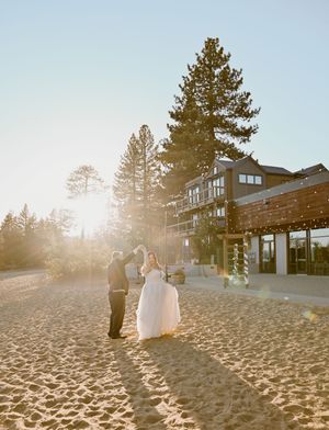 A wedding couple and their guests celebrate on a sandy beach as golden sunlight streams through tall pine trees at sunset.