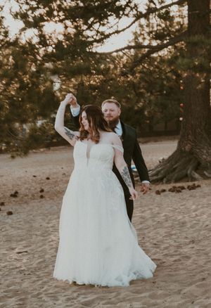 Couple in wedding attire dance and twirl together on a sandy beach at sunset with trees in the background.