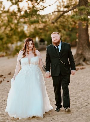 Wedding couple walking hand in hand along sandy beach under sunlit trees in full formal attire.