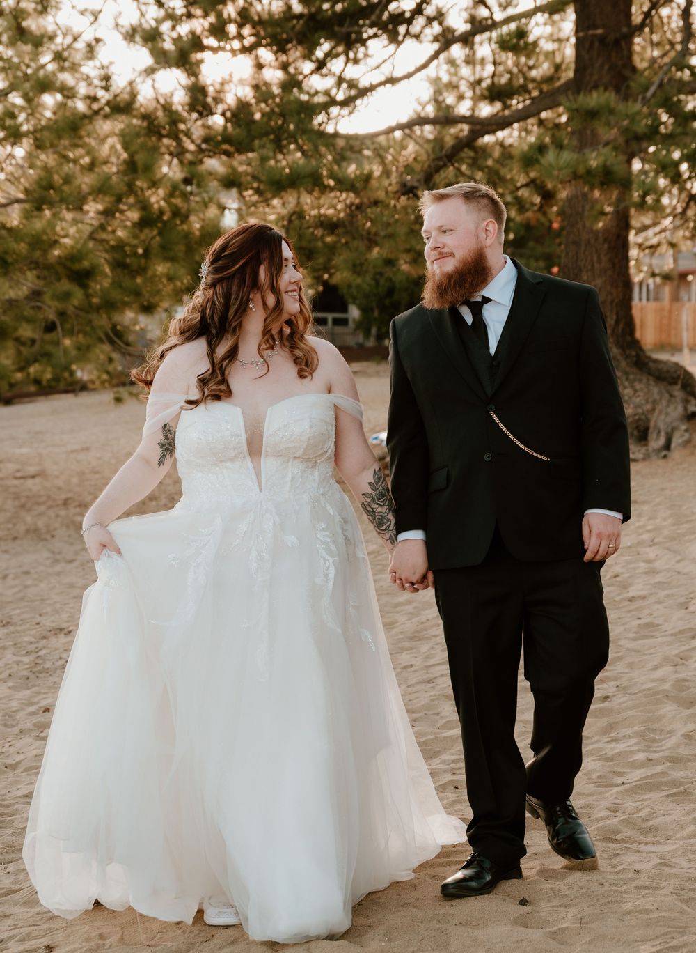A couple in wedding attire walks along sandy ground with sunset lighting, creating a romantic and intimate atmosphere.