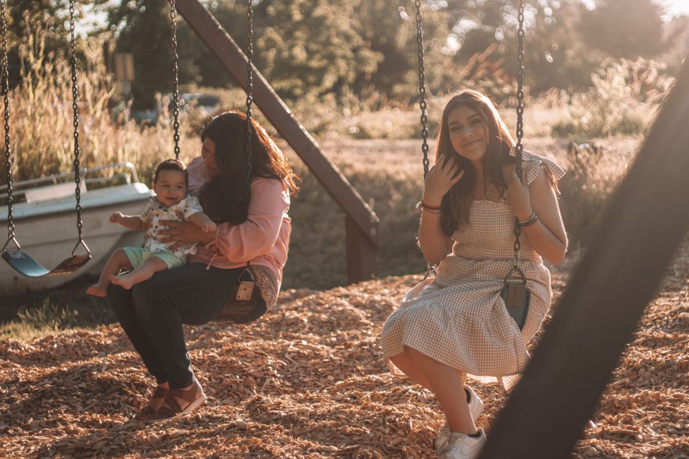 Two women sitting on swings at golden hour, one holding a baby in a sunlit outdoor setting.