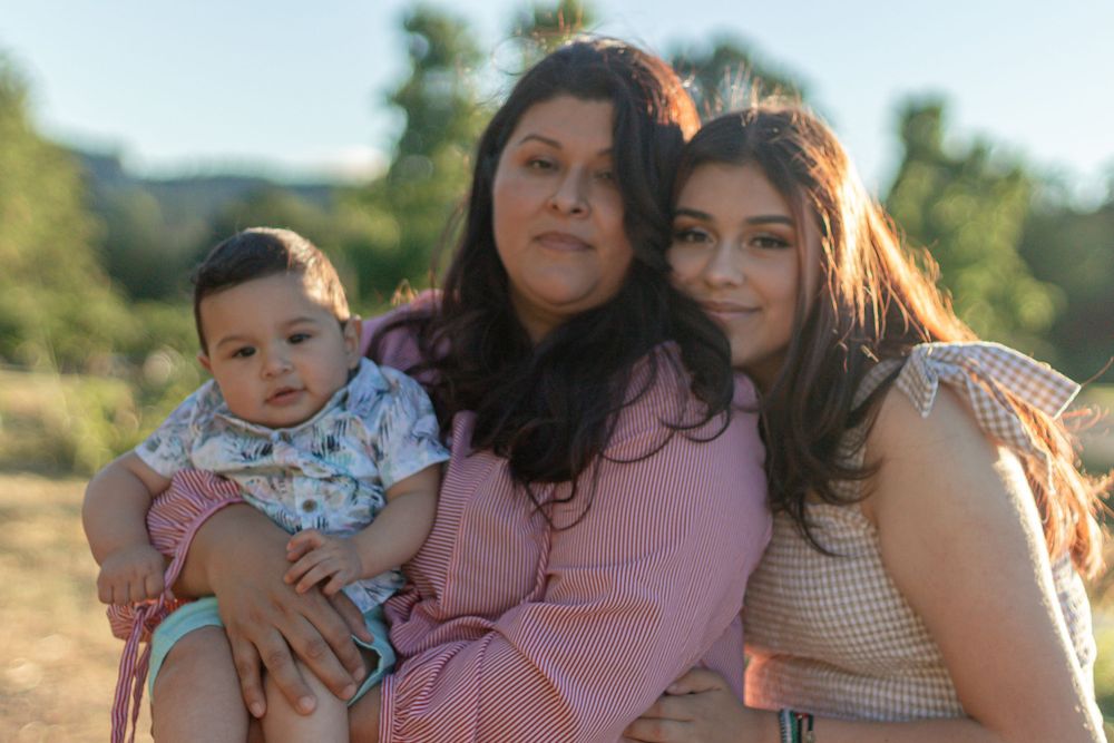 A woman with a baby and teenage girl pose together outdoors with green hills in the background.