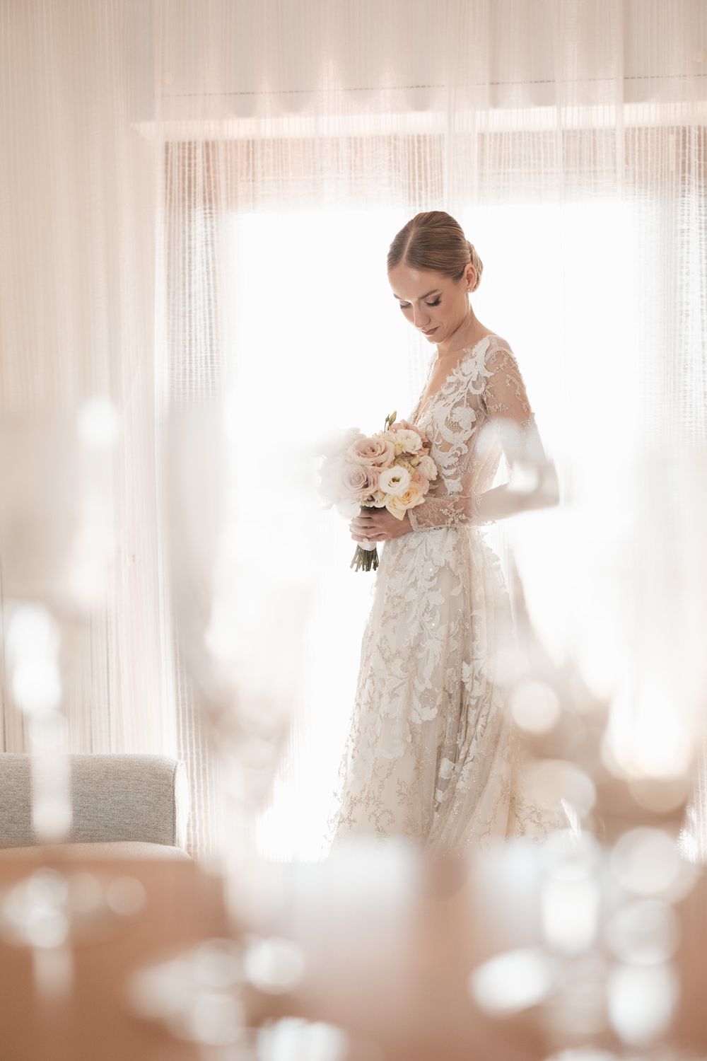 Elegant bridal portrait in lace gown holding bouquet against sheer white curtains.