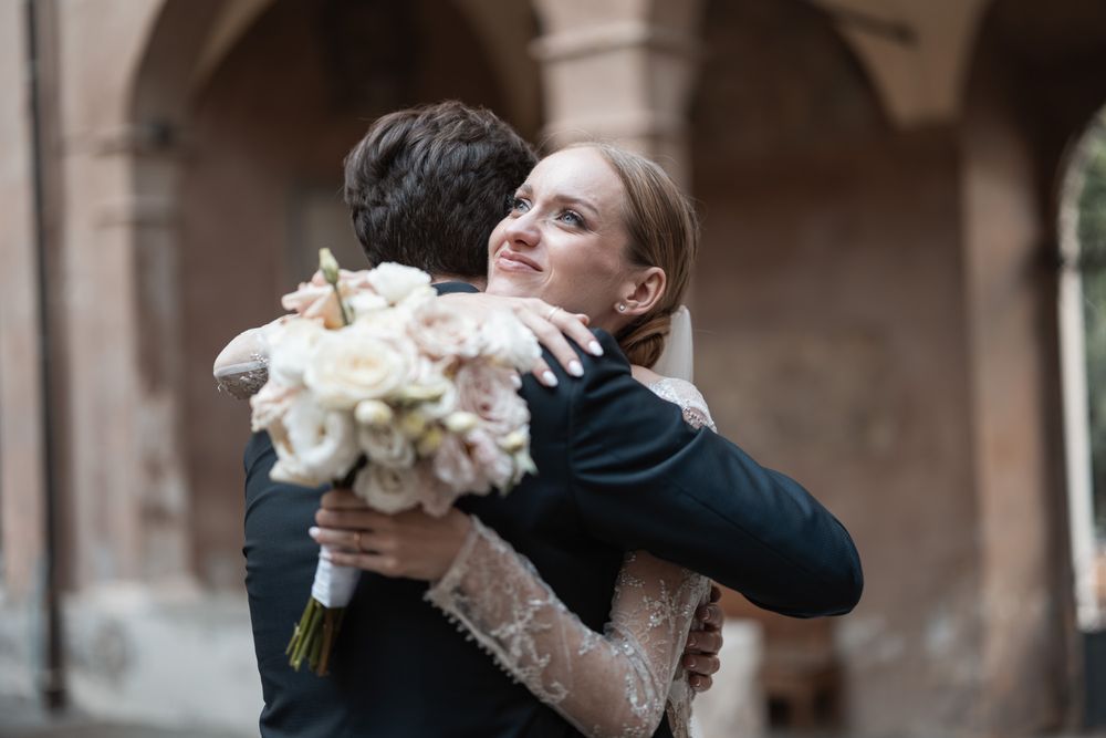 Tender embrace between newlyweds with white floral bouquet against arched architecture.