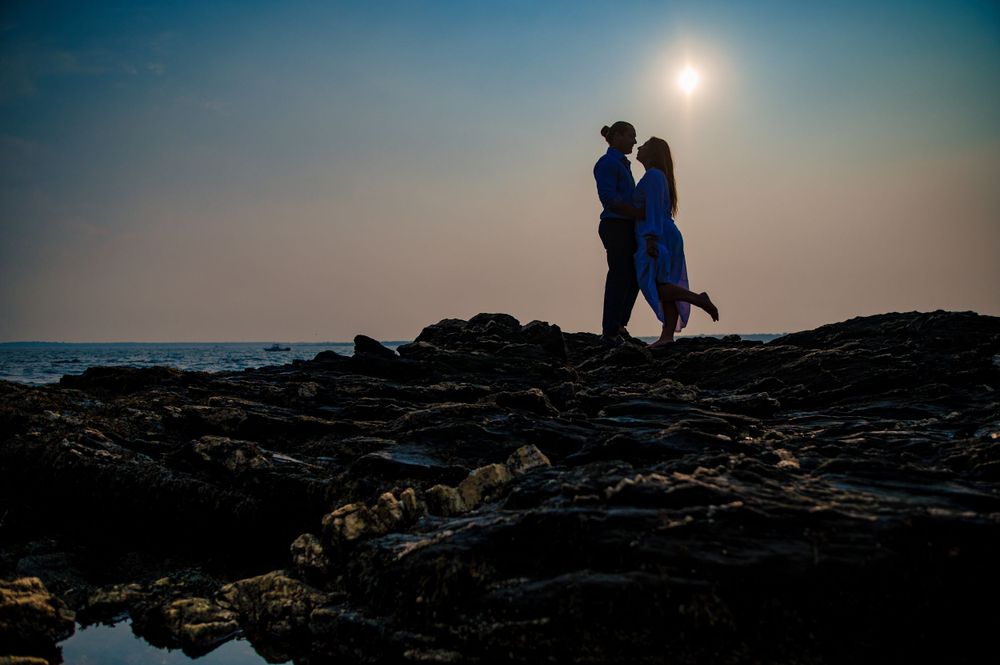 A romantic silhouette of a couple embracing on rocky coastal cliffs at sunset with glowing sun in background.