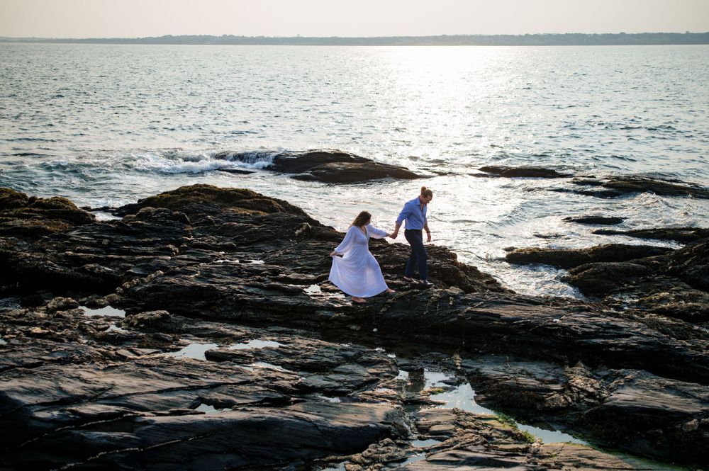 Two figures explore rocky coastal tide pools at sunset as waves crash against the rugged shoreline.