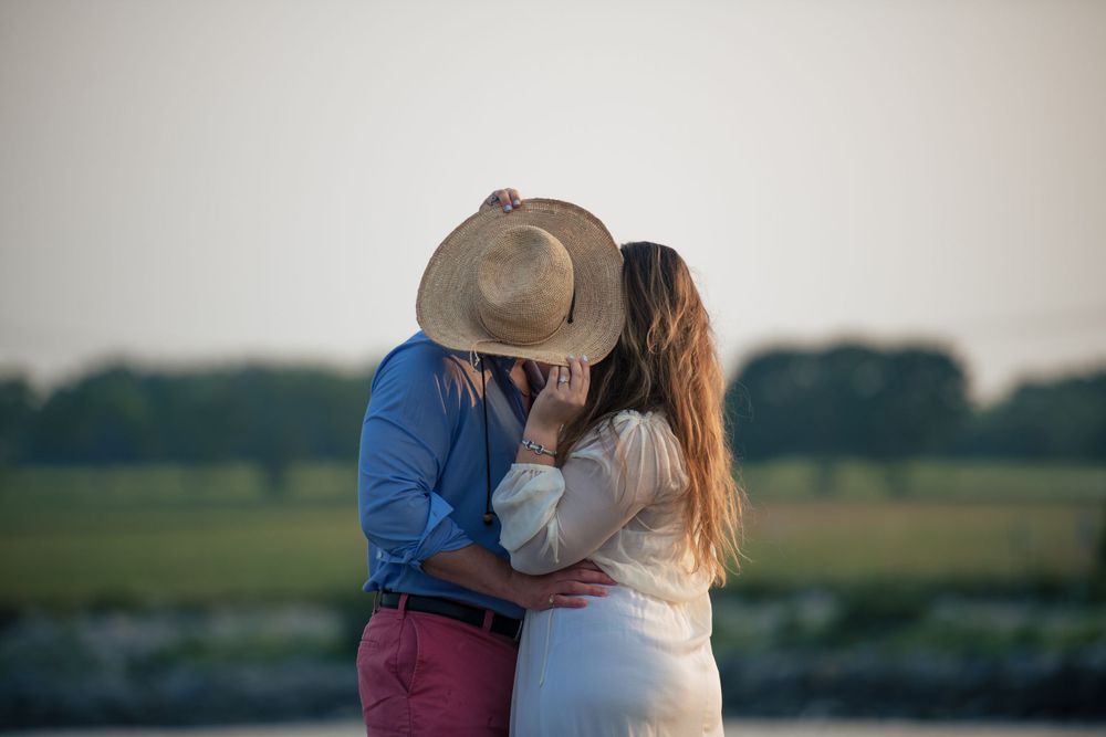 Two people share an intimate moment together at sunset in a rural field setting while wearing casual attire.