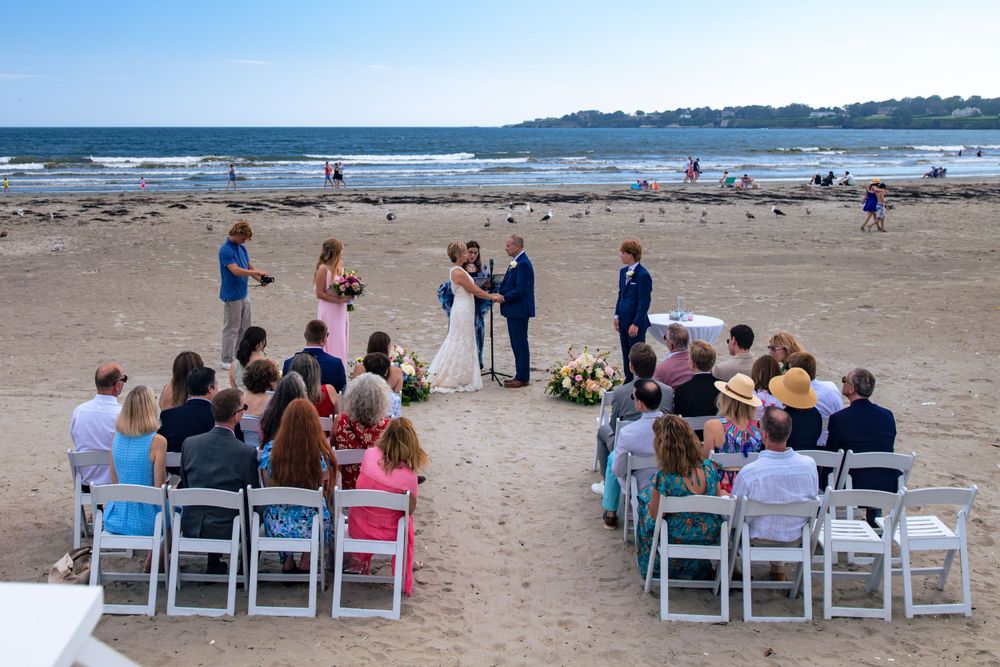 A small intimate beach wedding ceremony with white folding chairs set up on the sand near the ocean on a sunny day.