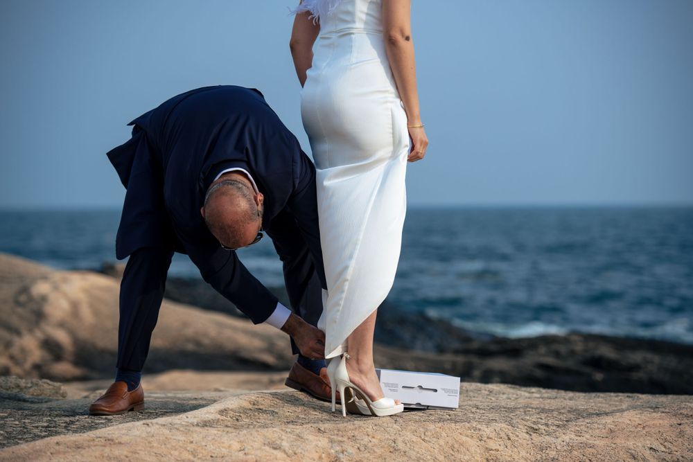 Two people on rocky coastal cliffs at sunset, with one in a white dress and another person bending down.