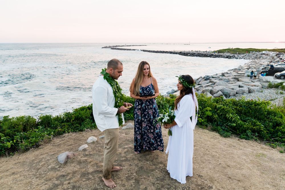 A beachside ceremony takes place at sunset with three people gathered on a cliff overlooking the rocky shoreline.