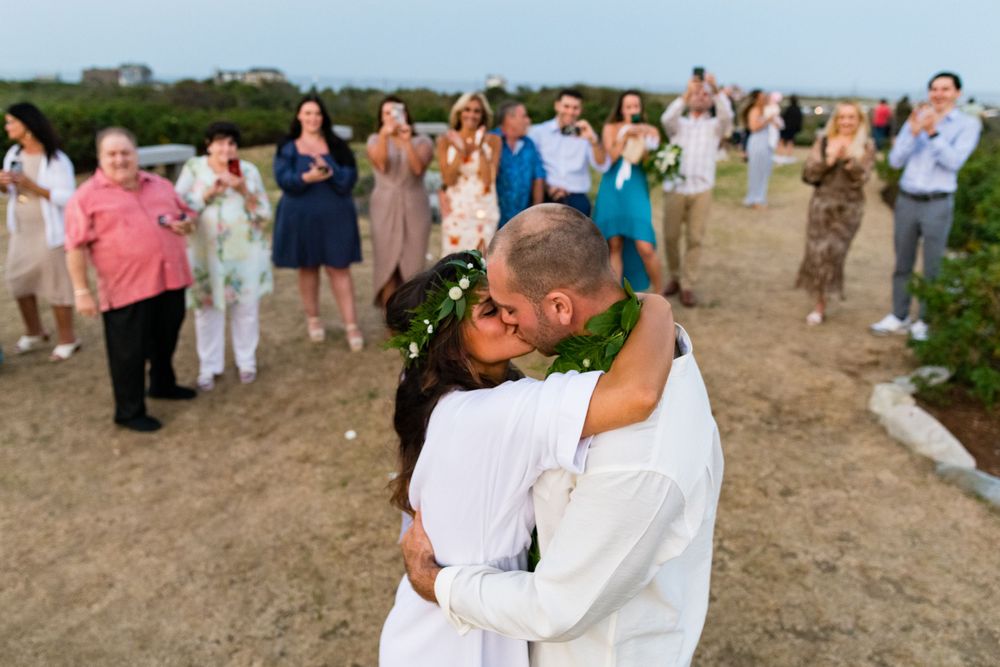Wedding guests photograph a couple's first kiss as newlyweds on a beach during their seaside celebration.