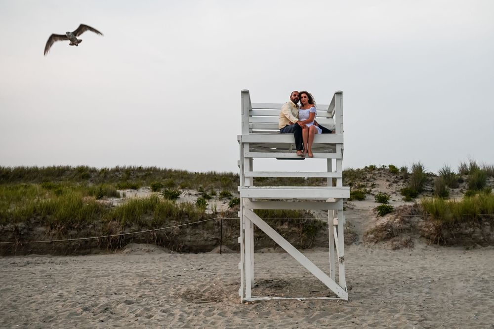 A white lifeguard chair stands on a beach with a seabird flying overhead at dusk.