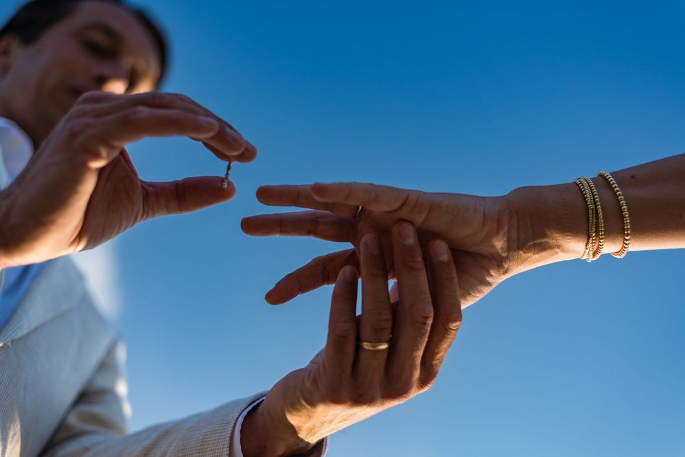 Groom places wedding ring on bride's finger against blue sky backdrop.
